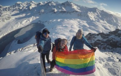 Three people on a snowy mountain top holding a rainbow flag